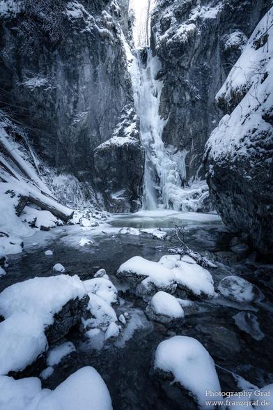 A stunning winter scene reveals a frozen waterfall cascading between towering, snow-covered cliffs. The waterfall, partially frozen in time, glistens with icicles that hang from the rocky ledges, creating a mesmerizing spectacle of nature's raw beauty. The cliffs, blanketed in a thick layer of snow and ice, frame the waterfall and add a sense of grandeur to the scene.

Below, a shallow stream flows gently over snow-dusted rocks, its surface partially covered with a thin layer of ice. The contra…