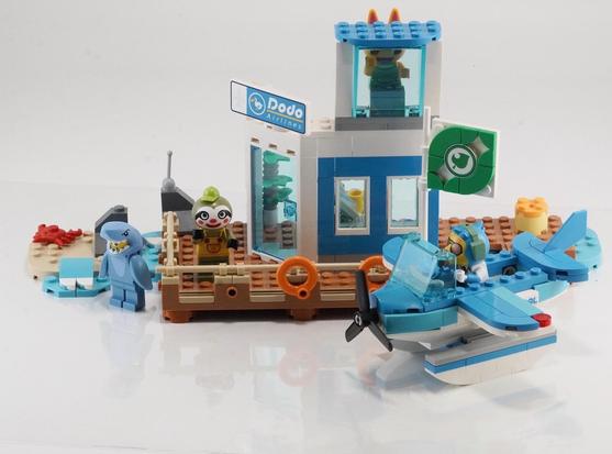 A colorful LEGO scene featuring a small airport building with a "Dodo Airlines" sign, an airplane, and three character figures: Lief, a shark, and a cat. The setting includes a dock, a beach area with a crab, and a Dodo in the seaplane. 