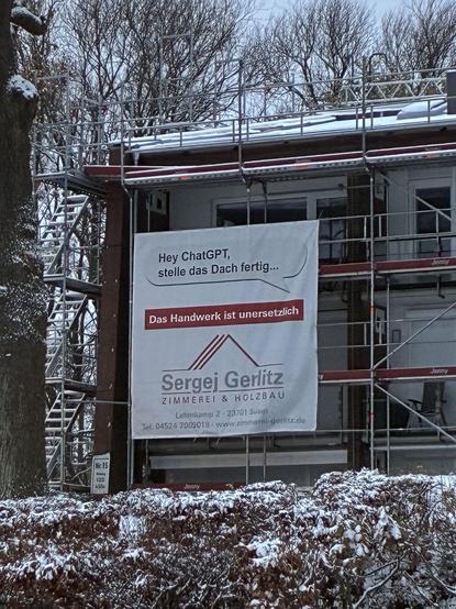 „Großes Werbebanner an einem verschneiten Baugerüst vor einem Haus. Auf dem Banner steht: ‚Hey ChatGPT, stelle das Dach fertig… Das Handwerk ist unersetzlich‘, darunter das Logo und die Kontaktdaten der Zimmerei Sergej Gerlitz. Im Vordergrund verschneite Büsche, im Hintergrund kahle Winterbäume.“
