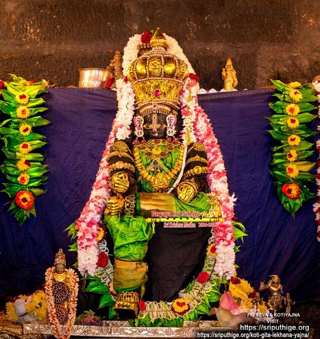 Idol of Hindu deity Krishna in bright green seated with right leg down, left leg folded flat, left foot placed over right knee, hands resting on respective legs; wearing gold shoes, gold palm coverings, an elaborate crested crown, and lots of gold ornaments. A string of red and white flowers around Krishna. A smaller string of white flowers around Krishna's (over the crown) ear-to-ear. A string of green betel leaves (red and yellow flowers on leaves) hangs in U shape in front the seat, Dark blue cloth background. A string of green betel leaves (red and yellow flowers on leaves) hangs down at each end of the background.