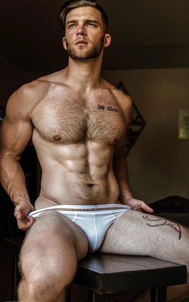 A muscular man with a "be nice" tattoo is sitting on a bench. He is wearing a pair of white briefs from Parke & Ronen brand