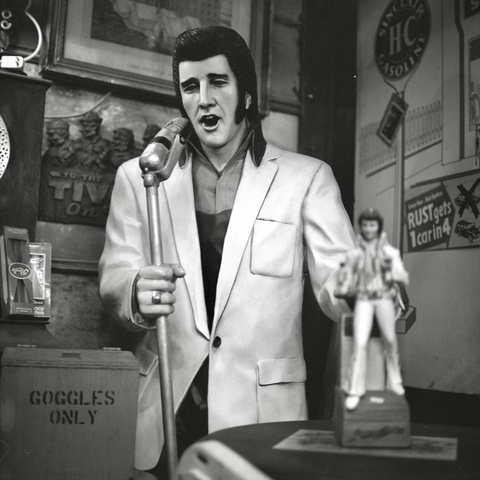 A black-and-white film photo. A life-sized statue of Elvis Presley, in his younger years, is holding a microphone and is singing, surrounded by random items in Broadway Paul’s Antiques & Salvage in Paige, Texas. Friday, November 7, 2025.