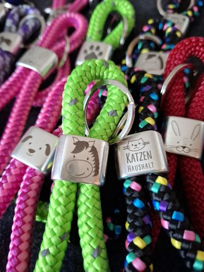 Mehrere bunte geflochtene Schlüsselanhänger mit silbernen Metallteilen, darauf gravierte Tiergesichter und die Worte 'KATZEN HAUSHALT'.
