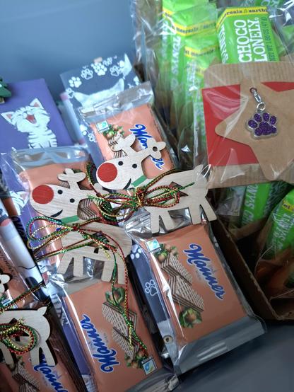 In Cellophan verpackte Süßigkeiten, im Vordergrund kleine Manner, auf Pappe geklebt, die mit Katzenmotiven überzogen sind. Drumherum rot-grün-goldene Schnüre mit Rentieranhängern aus Holz, daneben sieht man grüne Schokoladenriegel mit Kraftpapiertaschen, auf denen ein Pfotenanhänger angebracht ist.