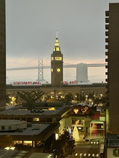 foggy morning in San Francisco. View of the San Francisco ferry building with the bay bridge behind it and a heavy layer of fog and a little bit of pinkish sunrise below it.