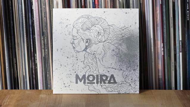 Ein künstlerisches Albumcover mit dem Titel „Moira“, das das Profil einer Frau mit wallendem Haar zeigt, dargestellt in einem monochromen, illustrierten Stil. Der Hintergrund ist mit Spritzern strukturiert, und das Cover ist auf einer Holzoberfläche vor einer LP-Sammlung platziert.