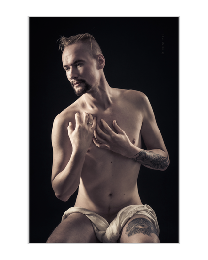 Artistic studio portrait of a seated man against a dark background, partially draped with light fabric around his hips. He poses with both hands on his chest in a sculptural, introspective gesture. Soft side lighting highlights his torso, facial expression, and tattoos on his arm and thigh, creating a dramatic and serene atmosphere. Photograph by Gus Martinie.