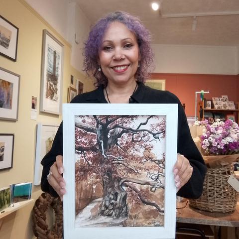 A woman with purple hair holds a painting of a twisty 500 year old Oak Tree  painted in earth pigments. Behind the woman is a gallery wall to the left and a bunch of dried flowers to the right.