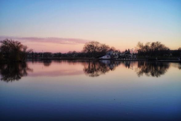 The calm water of a lake reflects the pale blue sky and the golden glow of the rising sun. Trees and some buildings can be seen on the distant shore in this soft warm image.