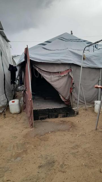 A makeshift shelter, likely a refugee tent, with a partially open flap revealing a dark interior, constructed of grey tarpaulin, positioned on a sandy, muddy ground, with other similar tents visible in the background.