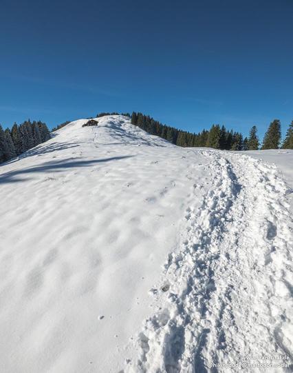 A stunning winter landscape unfolds under a clear, vibrant blue sky, capturing the pristine beauty of a snow-covered mountain slope. The untouched snow blankets the terrain, its smooth, white surface glistening in the bright sunlight. Footprints lead up the slope, creating a sense of adventure and exploration as they wind their way toward the summit.

At the top of the slope, a small, rustic cabin nestles comfortably among the snow-laden trees, its presence adding a touch of warmth and coziness…