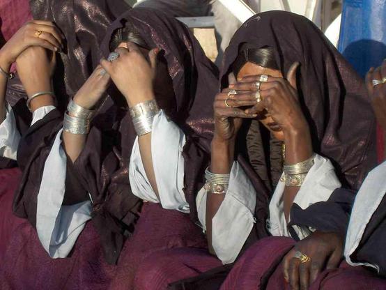 Several dark-skinned women draped in dark veils and white sleeves sit together, shielding their faces with hands adorned with multiple silver and gold rings and bracelets.