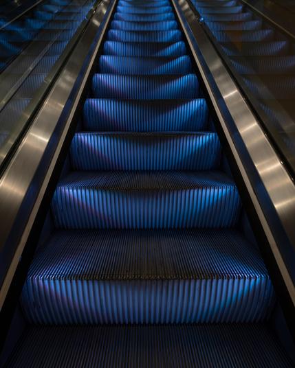 A photograph of the treads of an escalator, lit with blue light and framed by the railings. The treads are reflected in the glass of the railings. 
