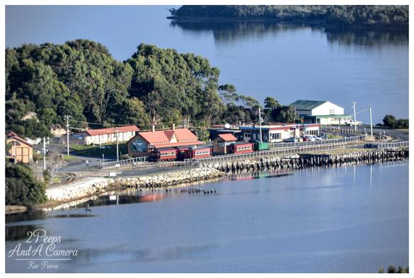 An elevated, distant shot of the Strahan train station complex nestled right on the edge of a calm, reflective body of water, likely Macquarie Harbour.

The station buildings feature terracotta roofs and a preserved red train is parked on the tracks.

The area is bordered by a rock sea wall and lush, dark green forest on the left, with other commercial buildings visible further along the waterfront road. The water is smooth and reflects the trees and distant landmass.