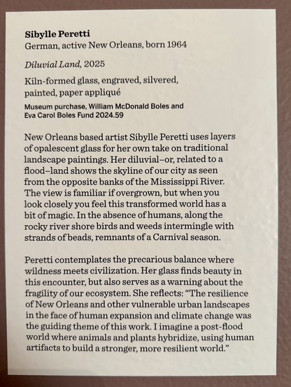 gallery sign: “Sibylle Peretti German, active New Orleans, born 1964 Diluvial Land, 2025 Kiln-formed glass, engraved, silvered, painted, paper appliqué Museum purchase, William McDonald Boles and Eva Carol Boles Fund 2024.59 New Orleans based artist Sibylle Peretti uses layers of opalescent glass for her own take on traditional landscape paintings. Her diluvial-or, related to a flood-land shows the skyline of our city as seen from the opposite banks of the Mississippi River. The view is familiar if overgrown, but when you look closely you feel this transformed world has a bit of magic. In the absence of humans, along the rocky river shore birds and weeds intermingle with strands of beads, remnants of a Carnival season. Peretti contemplates the precarious balance where wildness meets civilization. Her glass finds beauty in this encounter, but also serves as a warning about the fragility of our ecosystem. She reflects: "The resilience of New Orleans and other vulnerable urban landscapes in the face of human expansion and climate change was the guiding theme of this work. I imagine a post-flood world where animals and plants hybridize, using human artifacts to build a stronger, more resilient world."