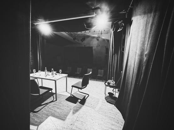 S/W-Bild von einem Hinterbühnenbereich auf die mit zwei Scheinwerfern beleuchtete Bühne. Dort ist neben einem kleinen Beistelltisch auch ein Esstisch mit Gläsern und Weinflasche sowie zwei Suhle zu sehen. Im Hintergrund sieht man im Halbdunkel den Publikumsbereich. 