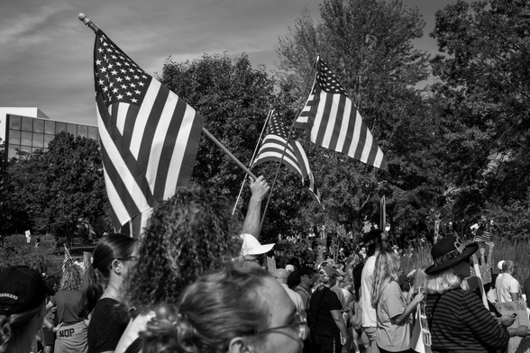 Black and white image of three US flags being held up over a crowd of people at a No Kings rally at the Iowa State Capitol in Des Moines, Iowa. 