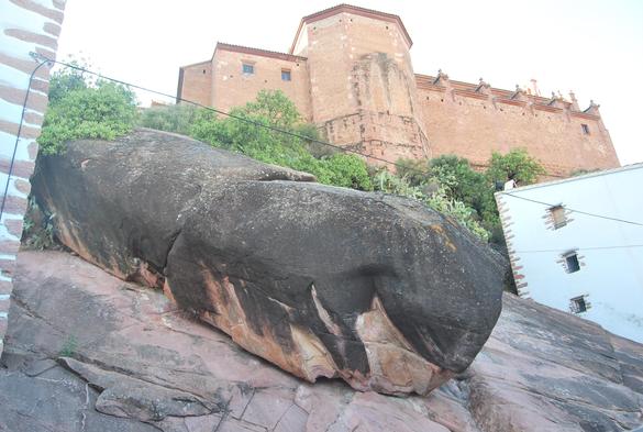 Roca larga y grande en una pendiente entre dos casas. Arriba, al fondo, se ve un castillo.
