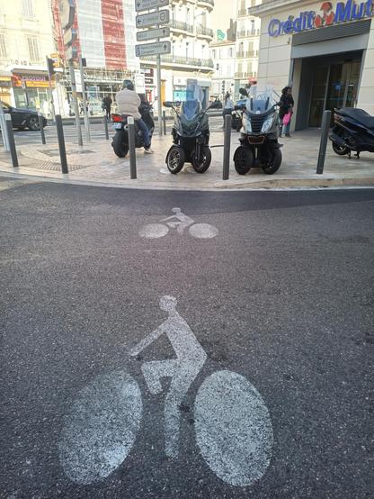 Photo d'une voie cyclable avec de slogo vélo peint au sol qui se continue sur un trottoir inaccessible car des scooters sont garés dessus. A gauche toujours sur le trottoir on aperçoit une motocycliste sur sa moto.