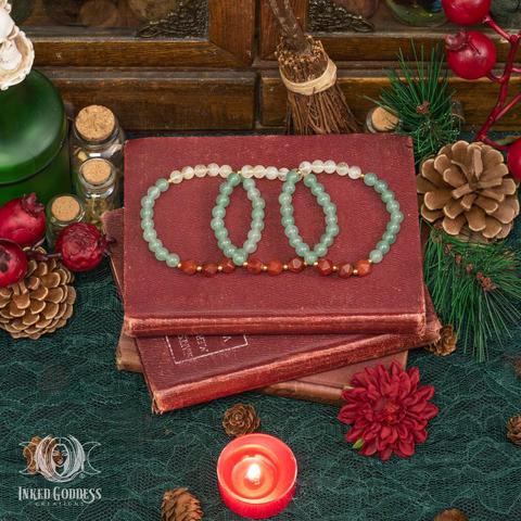 Three examples of the Yule Gemstone Bracelet Set sit atop a stack of small books. The bracelets are elastic and have no clasp. Each bracelet is made of small round Green Aventurine, Red Agate, and Citrine beads with smaller gold accent beads. This is an exclusive product from Inked Goddess Creations.