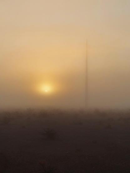 Photo of the rising sun just barely becoming visible through thick fog. The fuzzy silhouette of a tall, vertical spire rises up to the right of the sun.