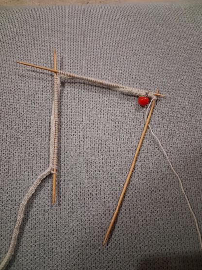 Photographie d'un tricot en cours. Un cordon libre s'attache à une première puis une deuxième et une troisième aiguille. Le cordon est de couleur beige clair.