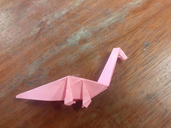 A pink origami plesiosaur style Nessie sitting on a wooden bench.