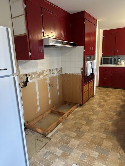 The West wall of our kitchen with the center cabinet holding our induction cooktop and all our pots and pans removed. A rectangle configuration of 2x4 boards is secured on the floor, and served as the base/toekick for it and the adjacent wall oven cabinet. Bare old tan Sheetrock from the 1950s is visible where the cabinet once resided. 