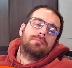 a tight frame of a man with a scruffy beard in a red-brown hoodie, we can only see his head, neck and top of shoulders. The man eyes almost entirely closed and are looking to his left. His head is tilted to the left and resting against the back of a shitty office chair that his company swears is fit for purpose.

He has glasses on, they are reflecting the horrible office florescent lighting, we can see one black headphone cord coming from his right ear and disappearing through the bottom of the frame.

The background is a couple shades of soul destroying grey that every office insists upon.