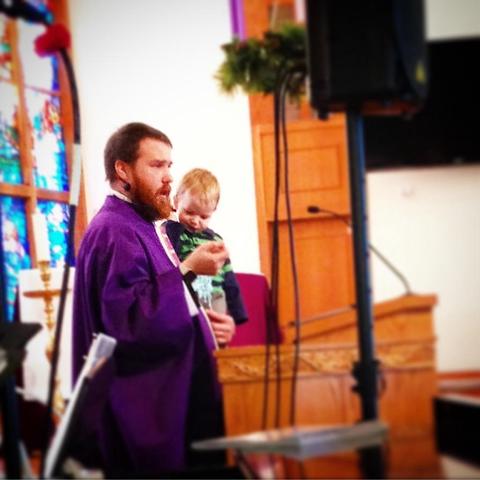 A man with a beard is speaking in front of a wooden lectern while holding a small child. The man is wearing a purple robe and appears to be in a place of worship, with colorful stained glass windows visible in the background.