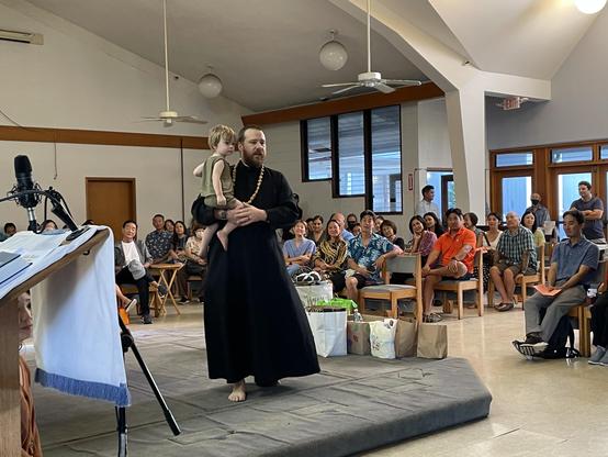 A man in traditional attire holds a young child while standing in front of an audience in a community setting. The audience appears engaged, seated on chairs with various expressions. There are items and bags on the floor, suggesting a gathering or event.