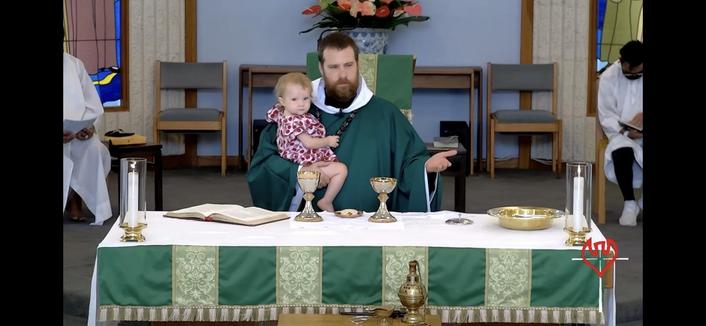A priest in green vestments is seated at an altar holding a toddler. The altar is decorated with lit candles, communion vessels, and a book. There are additional people in white robes in the background, and stained glass windows are visible.