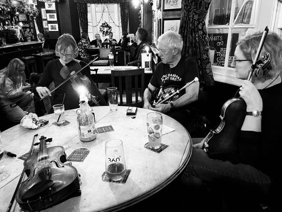 A fiddler and a piper playing while another fiddler listens, all around a round pub table with a candle and a fiddle on it.
