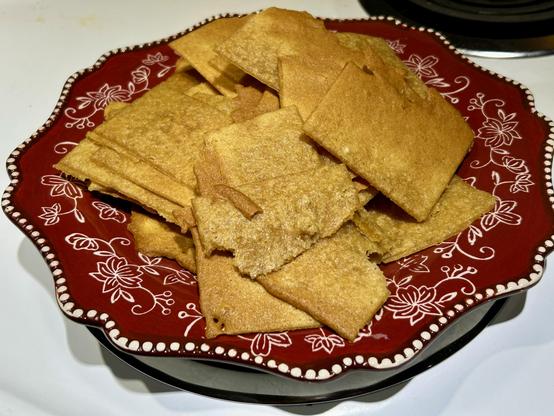 A pile of square and rectangular super thin gingerbread cookies atop a red plate with white floral patterns sitting on a white oven. One of the burners in the back is partially visible in the upper right corner.