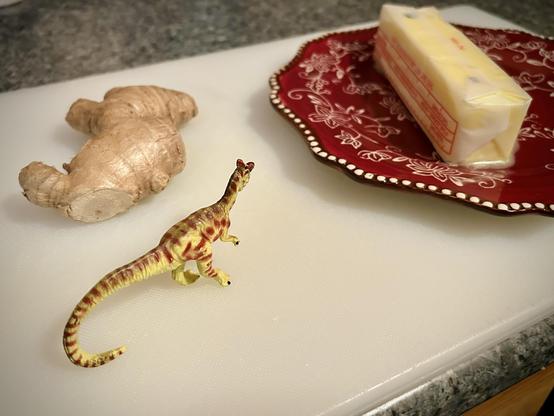 A green Dilophosaurus with brown spots stands on a cutting board with a fresh, unpeeled ginger root, and beside that a softened stick of butter on a red saucer with white floral patterns. The counter is a blue faux granite
