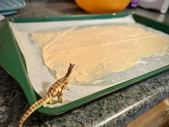 A green Dilophosaurus with brown spots stands on the edge of a green cookie sheet covered with a very thin layer of gingerbread batter waiting to go into the oven. The cookie sheet sits on a counter of blue faux granite. A large orange bowl is partially visible in the upper right corner. Sky blue pot holders are just in the frame of the upper left.