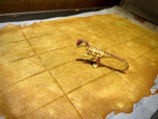A green Dilophosaurus with brown spots stands on a finished batch of gingerbread cookies atop a green cookie sheet that in turn sits atop a white oven.