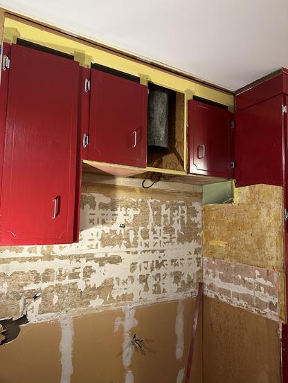 West wall of our kitchen, showing our red upper cabinets, and a scarred bald wall of 1850s old tan Sheetrock, where patches of old mastic cling to it, where off-white 6x6 porcelain tiles were once installed. They were also installed on the side of the oven cabinet to the right. 