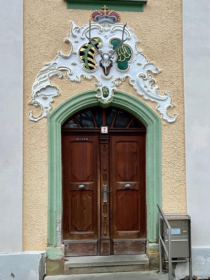 Eine Tür im lindgrün gestrichenen Bogen. Die Tür ist zweiflügelig und schließt mit einem Rundbogen, der fächerförmige Fensterchen hat, ab. Sie ist aus Holz und dunkel. In ihrer oberen Mitte trägt sie die Hausnummer 2 und links oben den Dreikönigs-Segen 20-CMB-25. Oberhalb der Tür verschnörkelter Stuck in weiß, obendrüber eine Krone in bunten Farben und darunter ein Geweih, das umgeben ist von Wappen. Ich kenne sie nicht, vermute links Thüriungen und rechts ? Die Hauswand ist gelb gestrichen, zwei Stufen führen zum Eingang. Rechts daneben ein Geländer aus Stahl sowie aus dem gleichen Material ein Briefkasten.