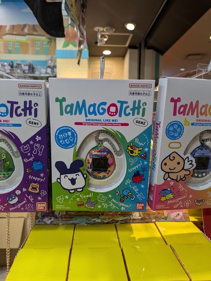 Three boxed Tamagotchi devices hanging on a store display. The center box is green and labeled “Gen2,” showing a pastel-speckled Tamagotchi inside. The boxes on either side are purple and pink, each featuring different character illustrations. All packaging has Bandai branding and colorful cartoon graphics.