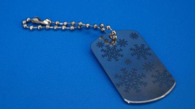 Nahaufnahme eines gravierten Edelstahl-Schlüsselanhängers mit mehreren Schneeflocken-Motiven. Der Anhänger liegt auf blauem Hintergrund, befestigt an einer silbernen Kugelkette.