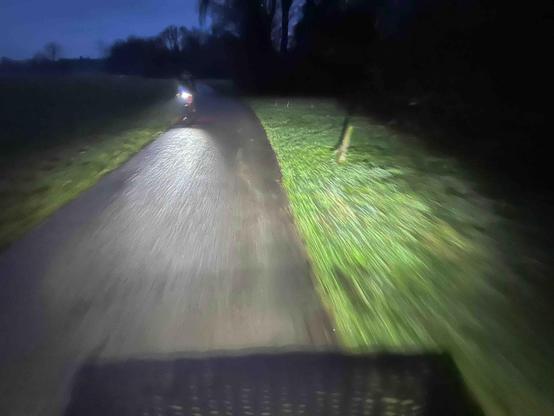 Ein einsamer, asphaltierter Feldweg an einem trüben, nassen, kalten Novembermorgen, aufgenommen vom fahrenden Fahrrad. Es ist noch nicht richtig hell, der Weg wird vom Fahrradlicht erhellt, in dem man den Schatten eines Lenkerkorbs erkennt. Auf der linken Wegseite sieht man den Scheinwerfer eines gerade entgegenkommenden Fahrrades