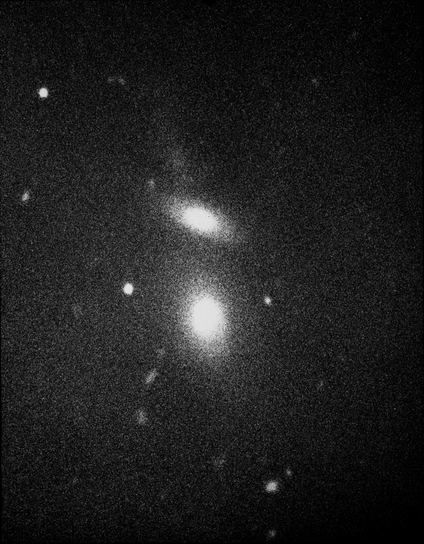 A grainy, black and white image of two galaxies. The bottom galaxy is roughly circular and the upper galaxy appears oval-shaped. Both have bright cores and become more translucent towards their edges. A fuzzy line appears above the top galaxy. A few white dots, which are stars, are seen on the black background.