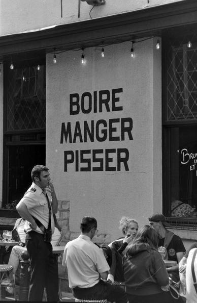 un vitrine ou l'on peut lirs "manger, boire, pisser" derriere une terrasse ou sont attablés des clients, et un homme debout en uniforme, perplexe