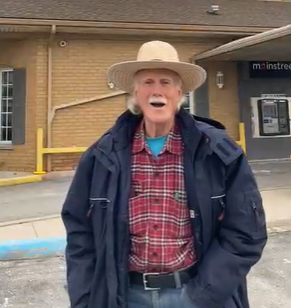 Tony standing outside the Mainstreet Community Credit Union. It is a modern  yellow brick building. Tony is dressed for the cool weather with his jacket open and is wearing a red, white and black plaid shirt with a bright blue T shirt visible and of course wearing his classic wide brimmed straw coloured working hat.