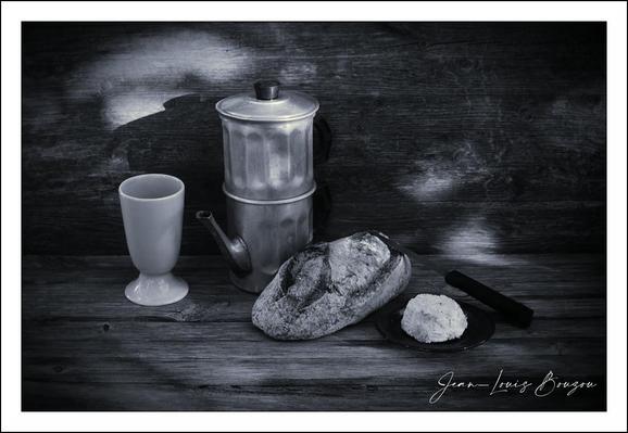 A quiet, contemplative still life rendered in rich black-and-white — the picture foregrounds simple objects on a weathered wooden plank: a metallic stovetop coffee maker (its brushed surface catching a soft highlight), a mazagran, a crusty loaf or roll dusted with flour, a round scoop of something soft (likely butter or cheese), and a dark-handled knife resting nearby. The photographer’s signature sits in the lower right corner, anchoring the work as a deliberate composition.
Light comes from the left, low and directional, carving the forms with crisp highlights and deep, velvety shadows. The moka pot’s cylindrical body and spout throw reflective gleams; its textured metal contrasts with the smooth, matte surface of the mazagran. The bread’s crust is tactile — cracked, irregular, with flour clinging to its ridges — while the soft ball beside it reads as a different material altogether, creamy and more diffuse in tone.
Textures dominate: the grain and scratches of the wooden tabletop and backdrop give a lived-in, rustic atmosphere; the contrast between hard metal, fragile ceramic, crisp crust, and soft spread creates a sensory dialogue about touch and taste. The composition feels carefully balanced — vertical weight from the coffee maker countered by the horizontal loaf, the knife providing a subtle diagonal that leads the eye through the scene.
