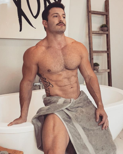 A topless muscular man with a towel around his waist is sitting on the edge of a bath in a bathroom