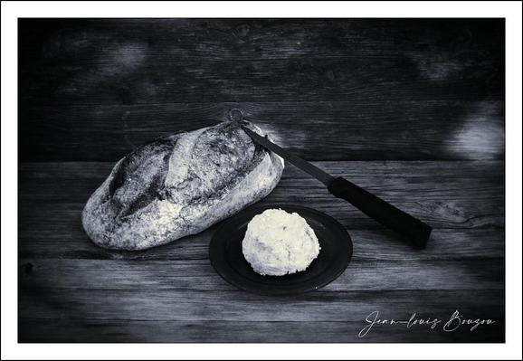 A quiet still life in high-contrast monochrome: a rustic, oval loaf of bread sits on weathered wooden planks, its crust scored with a single deep slash and dusted with a light veil of flour that catches the light. The crust is rugged and crackled, suggesting a hearty, hand-shaped boule; the interior hinting at soft crumb beneath the fissures. Beside it, a small dark plate holds a creamy cheese — smooth, softly peaked, with tiny textural ridges that imply softness and spreadability. A serrated knife rests above the plate, its blade angled into the scene as if just used and then set down, the handle pointing outward toward the viewer.
The composition uses negative space and directional lines well: the knife creates a gentle diagonal that leads the eye between bread and cheese, while the horizontal grain of the tabletop grounds the objects. The lighting is focused and directional, throwing subtle highlights across the flour and cream and deepening the shadows in the folds of the loaf, giving the image a tactile, almost audible quality — you can almost hear the crust break.
Artistically, the scene reads as a meditation on simplicity and sustenance. The monochrome palette strips away distraction, emphasizing texture and form and giving the image a timeless, almost nostalgic feel. Bread and cheese together are a universal symbol of nourishment and domestic ritual; the poised knife suggests a recent or imminent action, a human presence acknowledged only by the tools left behind.
