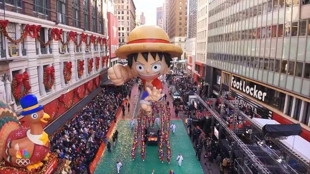 An overhead view of the Macy’s Thanksgiving Day Parade shows the enormous Luffy balloon towering above the street, with his straw hat, stretched-out punch pose, and cheerful expression clearly visible. Crowds line both sides of the green parade route, surrounded by festive holiday decorations. A large turkey float appears on the left, and buildings like Foot Locker frame the scene as parade handlers guide the balloon forward.