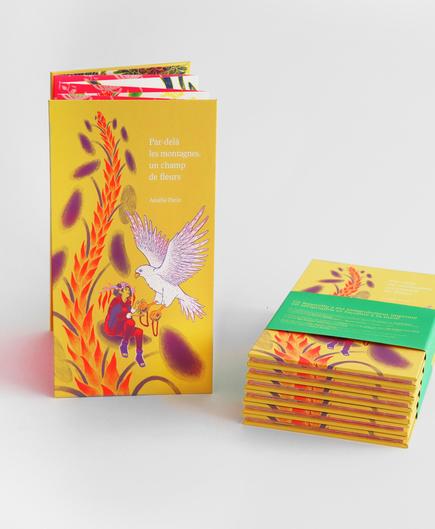Photographie d’un leporello intitulé « Par-delà les montagnes, un champ de fleurs », présenté debout, à côté d’une pile de plusieurs exemplaires fermés et maintenus par un bandeau vert. La couverture jaune montre une scène illustrée avec un faucon gerfaut en train de voler, tendant une énorme graine à une femme assise sur une feuille de pilée géante. Cette image est imprimée en sérigraphie 3 couleurs, blanc, rouge et violet, sur papier jaune. Le leporello a été imprimé en sérigraphie.